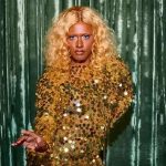 Drag queen com cabelo loiro encaracolado e vestido dourado brilhante.