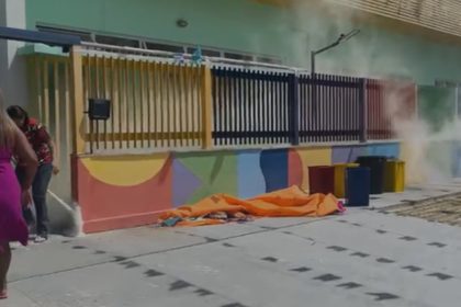 Princípio de incêndio na CMEI) Maria Emília Mestrinho, Duas mulheres perto de um prédio com fumaça, lixeiras e um pano laranja no chão.