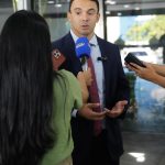 Entrevista coletiva com homem de terno, cercado por jornalistas com microfones e celulares.