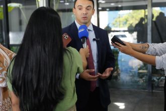 Entrevista coletiva com homem de terno, cercado por jornalistas com microfones e celulares.