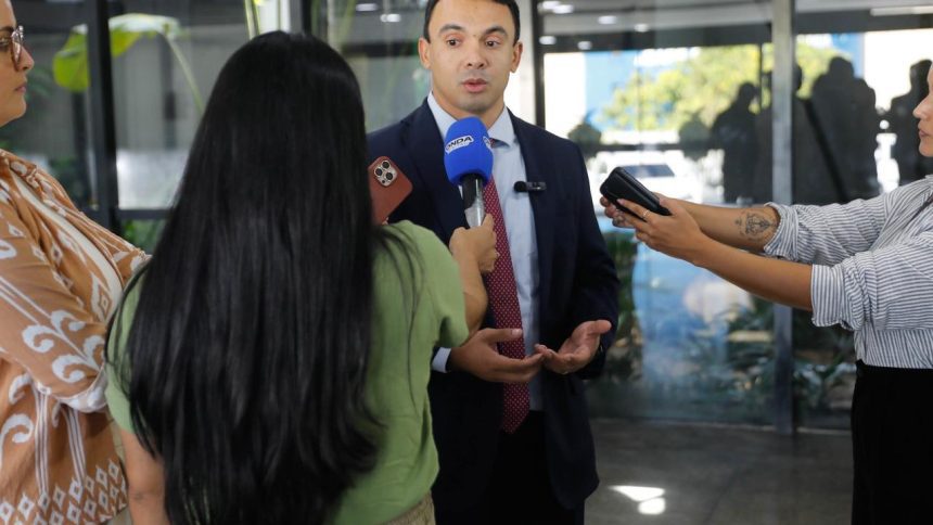 Entrevista coletiva com homem de terno, cercado por jornalistas com microfones e celulares.