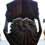 Estátuas segurando livros abertos do prêmio MEC, com foco em detalhes em bronze, simbolizando educação e conhecimento.