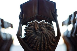 Estátuas segurando livros abertos do prêmio MEC, com foco em detalhes em bronze, simbolizando educação e conhecimento.