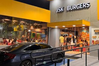 Fachada da lanchonete JSK Burgers com fila de clientes e carro estacionado em frente.