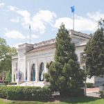 Fachada de um edifício com bandeiras, árvores e céu azul com nuvens.