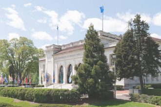 Fachada de um edifício com bandeiras, árvores e céu azul com nuvens.