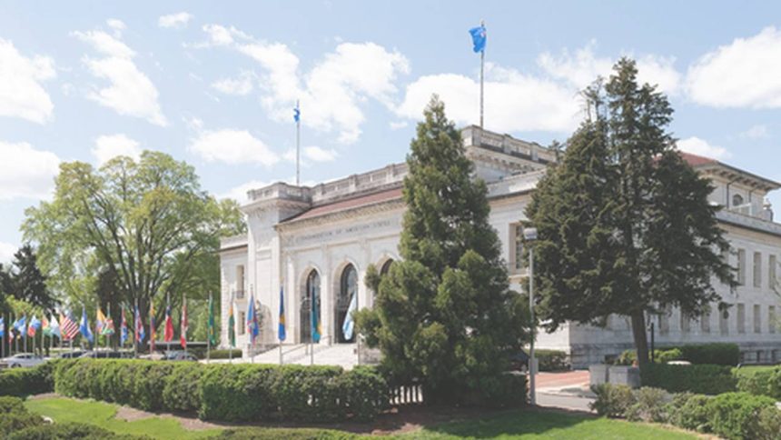 Fachada de um edifício com bandeiras, árvores e céu azul com nuvens.