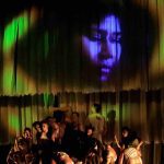 Grupo de dançarinos no palco com imagem de rosto projetada no fundo, em espetáculo de dança.