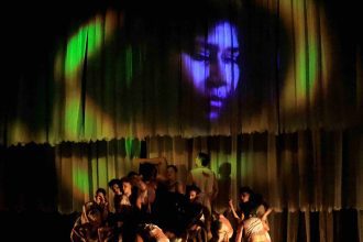 Grupo de dançarinos no palco com imagem de rosto projetada no fundo, em espetáculo de dança.