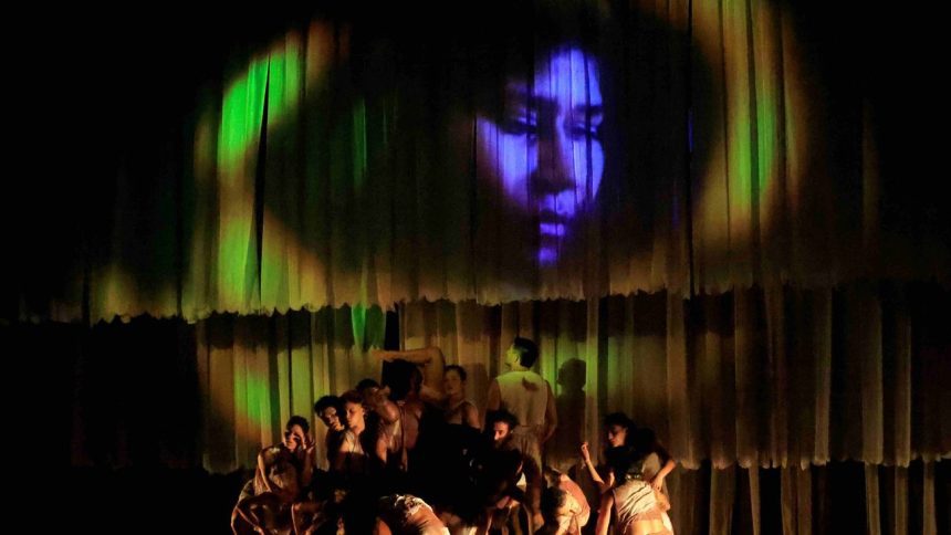Grupo de dançarinos no palco com imagem de rosto projetada no fundo, em espetáculo de dança.