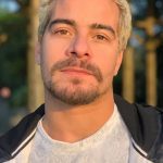 Homem com cabelo loiro e barba por fazer, usando jaqueta preta e camisa branca, em ambiente externo.