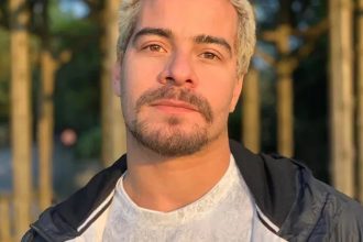 Homem com cabelo loiro e barba por fazer, usando jaqueta preta e camisa branca, em ambiente externo.