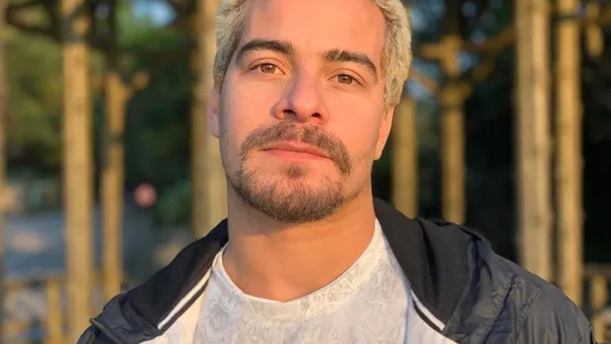 Homem com cabelo loiro e barba por fazer, usando jaqueta preta e camisa branca, em ambiente externo.