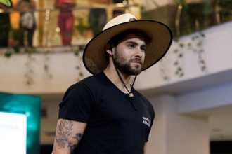 Homem com chapéu de palha e tatuagem no braço, possivelmente em um evento de moda.