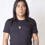 Homem de meia-idade com cabelo longo preto, camiseta preta e colar com pingente de pedra.