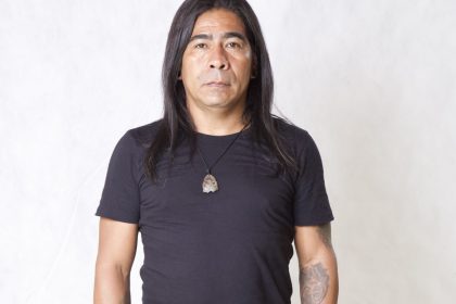 Homem de meia-idade com cabelo longo preto, camiseta preta e colar com pingente de pedra.