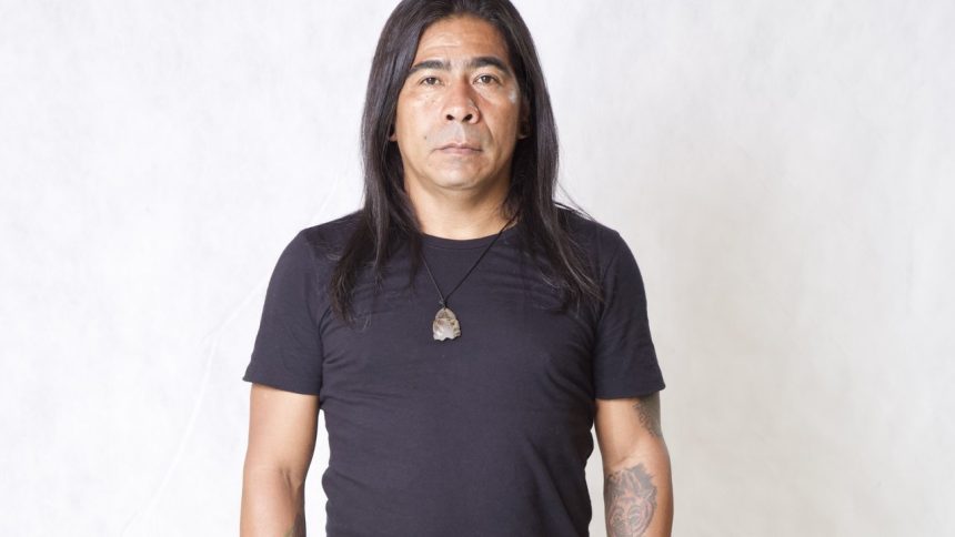 Homem de meia-idade com cabelo longo preto, camiseta preta e colar com pingente de pedra.