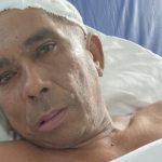 Homem em leito hospitalar com curativo na cabeça, possivelmente em busca de familiares.