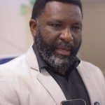 Reparação Homem negro de barba e terno fala, segurando um smartphone.