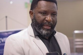 Reparação Homem negro de barba e terno fala, segurando um smartphone.