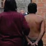 Homem preso com as mãos algemadas, acompanhado por policial