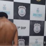 Homem preso em operação policial, encostado em uma parede com o brasão da polícia.