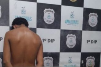 Homem preso em operação policial, encostado em uma parede com o brasão da polícia.