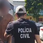 Homem preso sendo conduzido por policial civil. O homem está sem camisa, com tatuagens e algemado.