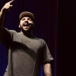 Homem sorrindo e apontando para cima em um palco, com roupa casual e boné.