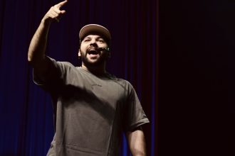 Homem sorrindo e apontando para cima em um palco, com roupa casual e boné.