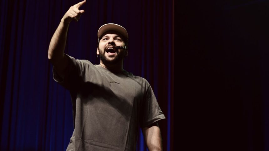 Homem sorrindo e apontando para cima em um palco, com roupa casual e boné.