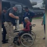 Idoso em cadeira de rodas sendo assistido por equipe de resgate perto de um helicóptero.