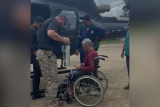 Idoso em cadeira de rodas sendo assistido por equipe de resgate perto de um helicóptero.