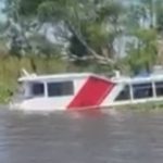 Lancha alagada em rio com parte submersa, com árvores ao fundo.