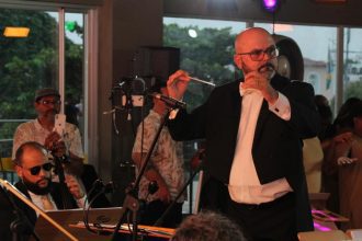 Maestro conduz orquestra, com músico tocando cello e bailarinos ao fundo.