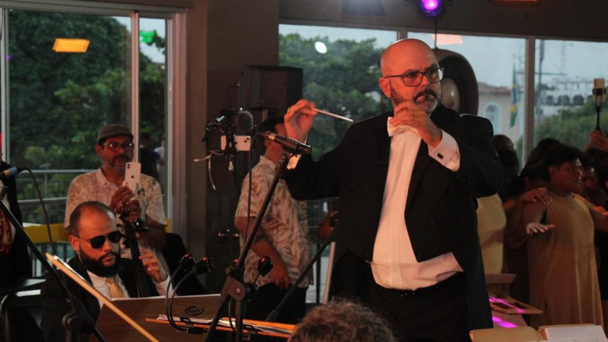 Maestro conduz orquestra, com músico tocando cello e bailarinos ao fundo.