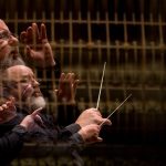 Maestro conduzindo orquestra, com efeito de múltiplas exposições, usando uma batuta e vestindo roupa preta.