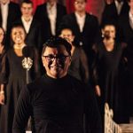 Maestro sorrindo com coral ao fundo.