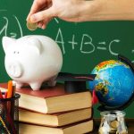 Mão depositando moeda em cofrinho de porco sobre livros, com globo e materiais escolares ao fundo, representando educação financeira.
