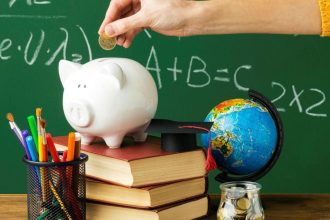 Mão depositando moeda em cofrinho de porco sobre livros, com globo e materiais escolares ao fundo, representando educação financeira.