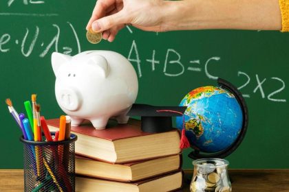 Mão depositando moeda em cofrinho de porco sobre livros, com globo e materiais escolares ao fundo, representando educação financeira.