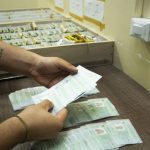 Mãos segurando documentos em frente a caixas, indicando possível registro ou distribuição.