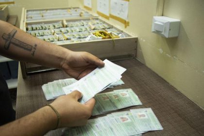 Mãos segurando documentos em frente a caixas, indicando possível registro ou distribuição.