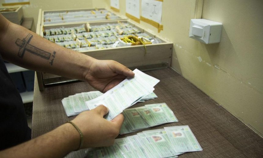 Mãos segurando documentos em frente a caixas, indicando possível registro ou distribuição.