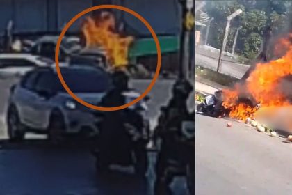Motocicleta em chamas após acidente trágico, com bombeiros e carro nas proximidades.