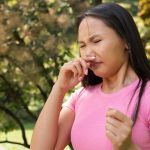 Mulher com alergia em um parque, coçando o nariz com expressão de desconforto.