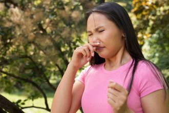 Mulher com alergia em um parque, coçando o nariz com expressão de desconforto.
