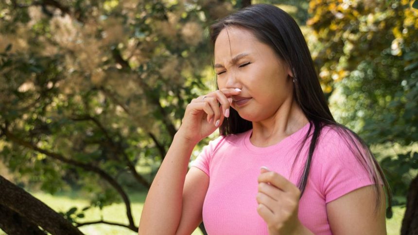 Mulher com alergia em um parque, coçando o nariz com expressão de desconforto.