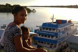 Mulher com criança em frente a um barco com a inscrição Previdência Social em um rio.