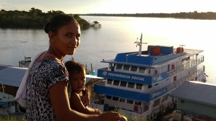 Mulher com criança em frente a um barco com a inscrição Previdência Social em um rio.